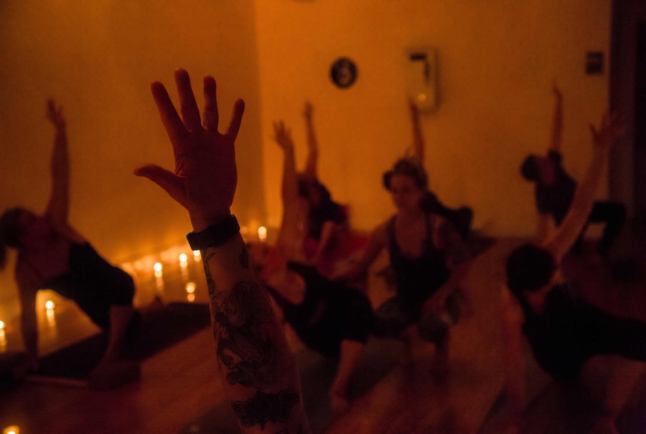 A tattooed hand raised in a candlelit movement practice — bodies in motion in the warm dark.