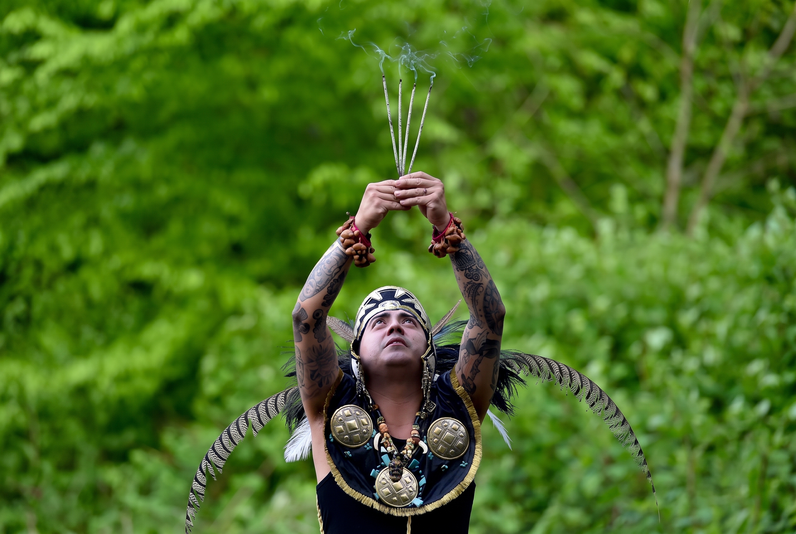 Smoking incense raised to the sky in ceremony — feathers and bronze medallions against a green forest.